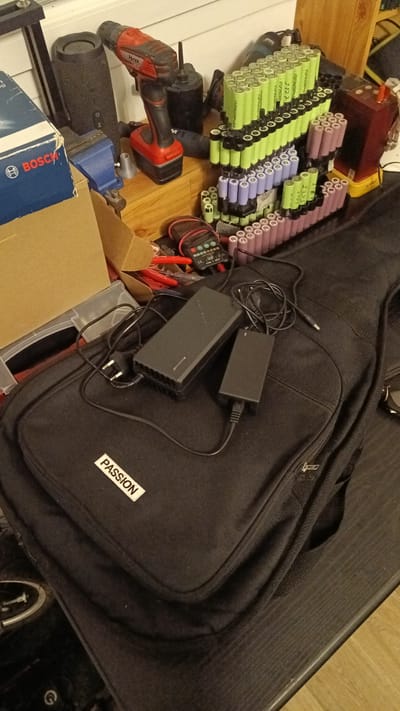 The image shows a collection of electronic components and tools on a wooden surface. In the foreground, a black fabric bag, possibly a guitar case, is partially visible with a white label that reads "PASSION." On top of the bag are two black power adapters, one larger than the other, connected by wires. In the background, a red and black power drill with a battery pack is prominent. To the left of the drill is a black cylindrical speaker. Further to the right, numerous cylindrical batteries, some green and some purple/pink, are stacked in plastic holders. A red and black digital multimeter and some red-handled tools are also visible amongst the batteries. A partially open brown cardboard box is to the left of the drill. The overall impression is of a workspace or storage area for electronic equipment. The image does not provide any information about the city of Villaz, France.