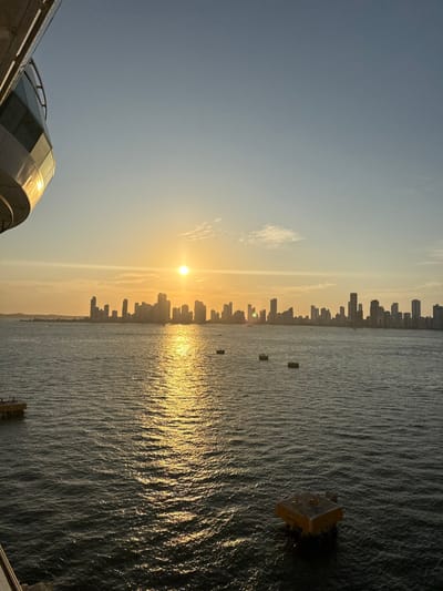 An expansive body of water stretches to a distant skyline illuminated by a bright, low sun. The sun's reflection shimmers on the water's surface, creating a golden path. A few objects float on the water's surface. The sky is a gradient of pale blue to a warm orange hue near the horizon. The lower portion of a structure, possibly a building, appears in the bottom left corner.
