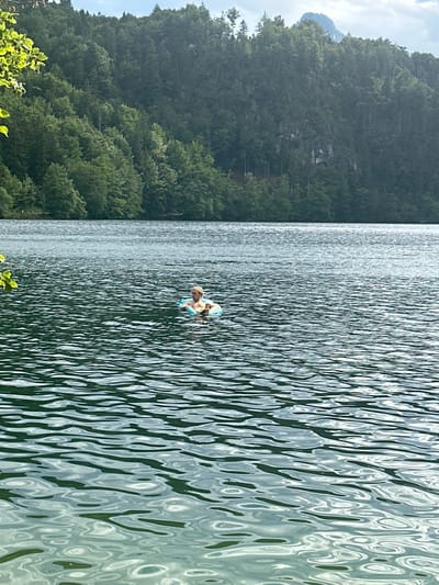 A person with blonde hair is floating in a blue inflatable ring in a body of water. The water is dark and appears wavy. In the background, a dense forest covers a hillside, with a peak visible above the trees. The scene is set in Kiefersfelden, Germany.