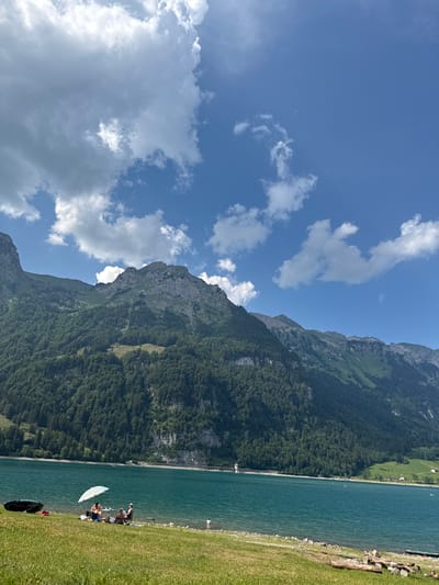 The image depicts a scenic view of a lake nestled beside a densely forested mountain range under a bright, partly cloudy blue sky. The scene is oriented vertically, with the sky occupying the top left and the lake and shoreline on the right. In the foreground on the right, a grassy slope leads down to the turquoise water.

Several people are visible on the grassy area by the water. Two individuals are seated in chairs near the lake's edge, and another person is lying down. A white, disc-shaped object, possibly a paddleboard or a large frisbee, rests on the grass near them. Further down the shoreline, a few other figures can be seen, and a dark object, potentially a backpack or a bag, is placed on the grass.

The mountain is covered in lush green trees, with rocky outcrops visible at its peak and along its slopes. The lake is calm and reflects the sky. A long, man-made structure, possibly a dam or a retaining wall, is visible along the far edge of the lake, separating it from the mountainous terrain. The overall impression is one of a sunny day, likely in the late morning or early afternoon, with people enjoying leisure time in a beautiful natural setting. There is no visible text in the image.
