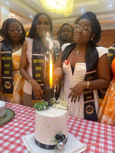 The image shows four women, all wearing black sashes with the words "Sisters for Life" and "Unity is Strength" written on them, gathered around a white cake decorated with a gold "Happy Anniversary" sign. A burning sparkler sits on top of the cake. The woman in the white dress is leaning in to blow out the sparkler. She is wearing a gold chain necklace and a gold bracelet. The woman behind her is wearing sunglasses, has a black sash over her pink blouse, and is holding the sparkler with her left hand. The woman on the left is wearing an orange and white dress and is also wearing a black sash. The fourth woman is wearing an orange and white dress and is only partially visible. The table has a red and white checkered tablecloth. The scene takes place inside a restaurant in Buea, Cameroon, located at coordinates 4.145, 9.23. The walls of the restaurant are a warm yellow color, and there is a decorative chandelier hanging from the ceiling. The overall atmosphere is festive and celebratory.