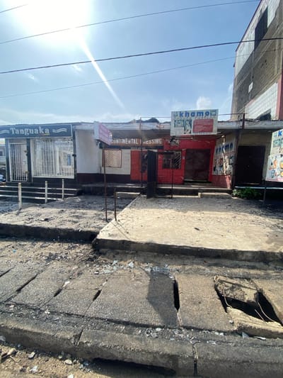 The image shows a street scene in Bonadikombo, Cameroon, located at 4.048° N, 9.218° E. The street is lined with small, one-story buildings. The image is taken from the street looking toward the buildings. The closest building is a shop with a sign that reads "Continental Pressing". It has a red brick facade and a metal gate in front. The shop next door is a beauty salon called "Khama" with a blue sign. The building on the left has a sign that reads "Tanguaka". There is a concrete sidewalk in front of the shops with some trash and debris scattered around. The sky is blue with some white clouds. The sun is shining brightly in the sky, casting a glare on the image. The buildings are all in relatively good condition, but there is some damage to the sidewalk, suggesting a recent event. The image suggests a busy commercial street with numerous small businesses. The image was taken in broad daylight.