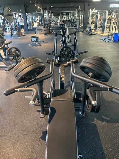 This image depicts the interior of a fitness gym located in Jestetten, Germany. In the immediate foreground, a black, plate-loaded chest press or shoulder press machine is viewed from behind its seat, prominently featuring multiple circular black weight plates stacked on each side of its lever arms. The machine bears a "Technogym" logo. The gym floor is covered with dark, interlocking rubber mats.
In the mid-ground and background, various other black strength training machines, benches, and weight racks are visible. Several individuals are present: one male is standing near a weight rack on the left, wearing a light t-shirt and dark shorts, facing away from the camera. Another male is partially visible, seated on a machine on the right. Additional figures and equipment are reflected in the large mirrors lining the back wall. Overhead lighting fixtures illuminate the space. On the far right wall, a neon sign reads "STRENGTH," and a blue bin sits next to a coiled rope on the floor.