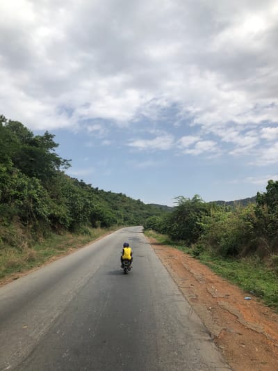 The image shows a motorcyclist wearing a yellow shirt and blue helmet riding down a paved road in Assob Hausa, Nigeria. The road stretches into the distance and is flanked on both sides by green vegetation. The sky is partly cloudy. The adjacent ground is brown soil and vegetation, with some trash visible.