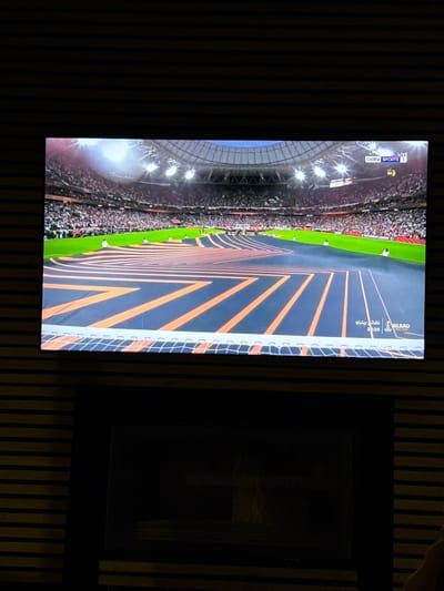 The image shows a flat-screen television displaying a live broadcast of a sporting event in a stadium. The stadium is filled with spectators, the field is empty, and the broadcast is showing a view of the field from above. The broadcast is for a sports channel called beIN SPORTS 1, and the television is mounted on a wall in a room with wood paneling. The coordinates 32.689, 35.054 locate the city of Daliyat al-Karmel, Israel.