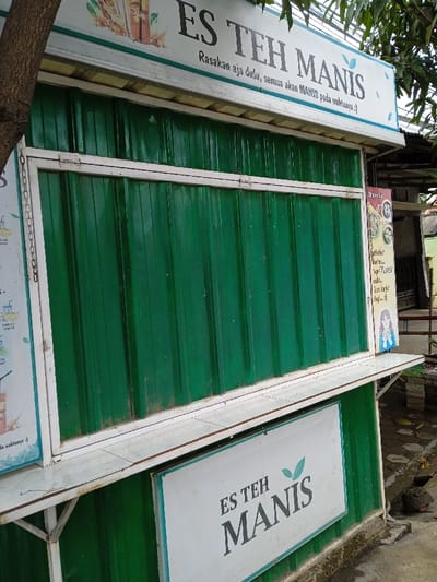 The image shows a closed "Es Teh Manis" (Sweet Ice Tea) stall. The stall is green with white trim and signage. One sign reads "Es Teh Manis" with the slogan "Rasakan aja dulu, semua akan MANIS pada waktunya :)" ("Taste it first, everything will be SWEET in its time :)"). Another sign has the words "Es Teh Manis" on it. A menu is partially visible on the side of the stall. The stall appears to be constructed of corrugated metal.