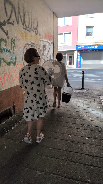 Two women are in a covered walkway with a cobblestone ground and a concrete wall covered in graffiti on the left. The woman in the foreground is wearing a white dress with black floral patterns and white sandals. She is looking up and to her left. To her right, a large white fan is positioned on the ground. Behind her, a second woman in a white dress and white flip-flops is holding a black bag and looking towards the fan. A street lamp is visible in the background. The scene appears to be set during the daytime with bright sunlight casting shadows. A blue sign with white text, "spielhalle EINGANG", is visible on a building in the distance. The overall mood is casual, possibly a warm day where the fan is being used for cooling. The setting suggests an urban environment in Germany, consistent with Osnabrück.