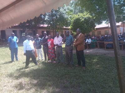 The image depicts an outdoor gathering in Idakho South ward, Kenya, under natural daylight. In the foreground, a light-colored tent canopy partially shades a grassy area where a group of ten adults stands. From left to right, individuals are dressed in varied attire including blue and patterned shirts, a light blue shirt, a plaid shirt, dark polo, red patterned dress, striped shirts, and a brown blazer. One man in a green striped polo holds a microphone, while a man in a brown blazer faces him attentively. In the mid-ground, a large audience of approximately 50-100 adults is seated on benches, observing the foreground activity. Behind the audience, single-story light-colored buildings with reddish corrugated iron roofs are visible, surrounded by green trees. Shadows on the grass indicate bright sunlight.