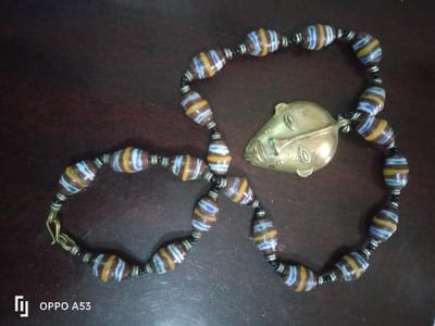 A handcrafted necklace is displayed on a dark brown, textured surface, specifically located in Riabai ward, Kenya. The necklace consists of numerous elongated, barrel-shaped beads, each featuring horizontal stripes in brown, white, yellow, and blue. These larger beads are separated by smaller, dark-colored spacer beads. A brass-toned metal pendant, shaped as a stylized human face with prominent nose and eyes, is centrally attached. One end of the necklace shows a brass-colored hook-style clasp. No individuals are present. The image was captured using an OPPO A53 device, as indicated by the watermark.