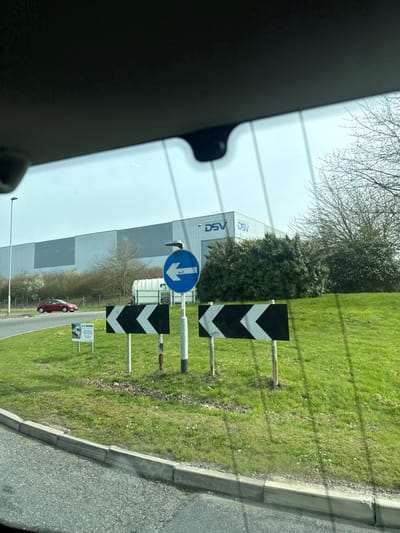 The image, taken from within a vehicle, captures a street scene at a roundabout in Thrapston, United Kingdom, specifically near a DSV logistics facility. The foreground shows the interior top edge of a vehicle's windshield, including a rearview mirror housing and a dashcam or sensor unit. Parallel lines, possibly defrosting elements, are reflected on the windshield.
The scene focuses on the central grassy mound of a roundabout, covered in short green grass with scattered small white flowers. Three traffic signs are centrally positioned on this mound: two rectangular black signs with white left-pointing chevrons, and one circular blue sign with a white left-pointing arrow mounted above them on a taller pole. To the left of these signs, a smaller white sign with black text, partially visible, displays "A C BOWNS" and contact details, likely an advertisement. Behind the signs, a white skip or trailer with a green lid is visible on the mound.
An asphalt road surface with a concrete kerb defines the edge of the roundabout. On the far side of the roundabout, a red hatchback car, possibly a Mazda model, is observed moving right-to-left. In the background, a large, light grey industrial building with the "DSV" logo displayed prominently multiple times on its facade is visible. The area around the building and the roundabout is bordered by various trees and bushes, some with bare branches and others with green foliage. A street light pole stands on the far left. The sky is clear and bright. No individuals are visible in the frame.