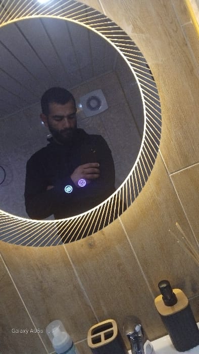 An individual with dark hair and a beard, wearing a dark long-sleeved garment, is depicted in a mirror selfie taken within a private residence in Oreshene, Bulgaria. The subject holds a smartphone, which reflects two illuminated icons (one blue grid pattern, one purple power symbol) onto their left sleeve. The mirror is circular with an illuminated border featuring a diagonal linear design. The reflection shows a light-tiled wall with a white exhaust fan positioned behind the subject's head. A paneled ceiling is also visible in the reflection. Below the mirror, on a white sink counter, are a white spray bottle, a dark and light-brown container resembling a toothbrush holder, a metallic faucet, and a dark and light-brown soap dispenser. A reed diffuser is partially visible on the right. The walls surrounding the mirror and counter are clad in light brown rectangular tiles. The image bears a "Galaxy A05s" watermark in the lower-left corner.