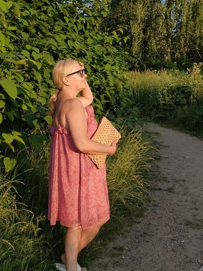 This media file captures a woman standing on a dirt path next to lush green foliage. She is looking off to her right, with her right hand resting on her forehead, shielding her eyes from the sun. She is wearing a pink, patterned dress and black-rimmed glasses, and she is holding a woven clutch bag. The scene appears to be set in a natural environment, possibly a park or a nature reserve, with trees and tall grass visible in the background. The sunlight suggests it is daytime, likely late afternoon due to the warm lighting and long shadows. The woman's posture and hand gesture might indicate she is observing something in the distance or feeling the warmth of the sun. There is no visible text in the image. The location context provided suggests the scene is at the Tourist Pension "Klyazminskoye Reservoir" in Russia.