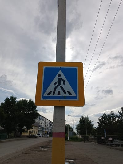 The image shows a pedestrian crossing sign mounted on a concrete pole. The sign is square with a yellow border and a blue interior, depicting a black silhouette of a person walking across white stripes. The pole itself is painted with yellow and red stripes at its base. In the background, there is a street with parked cars and a multi-story apartment building. Trees line the street and the sidewalk. The sky is cloudy. The location is identified as being in the city of Atbasar, Kazakhstan. The scene appears to be an ordinary urban street setting.
