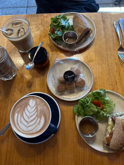 A wooden table displays two plates, each with a halved sandwich and small salad, and a metal ramekin. A clear jar of small cookies sits between them. A glass of iced coffee with a metal straw and a closed jar with a spoon are also present. A dark blue cup and saucer hold a latte with latte art, with a spoon nearby. Silverware and napkins are seen.