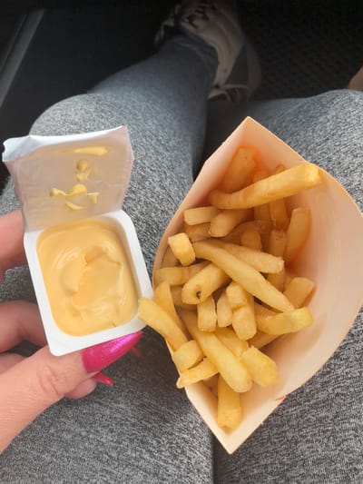 A person is holding a container of cheese sauce and a container of french fries in their lap. The person is wearing gray sweatpants and gray sneakers. The photograph was taken in an indoor setting, possibly a car. It is not possible to determine if the location is in the city of Kstovo, Russia from the image alone.