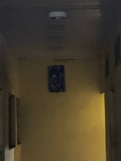 The image depicts an interior space with a wall featuring an electrical box opening. The room appears to be illuminated by overhead lighting, casting a yellow tint. In the foreground, what seems to be steps or tiered surfaces are visible. On the left side of the frame, a gray wall is present, and a circular object is mounted on it. There is no evidence of people, actions, or interactions. The image's location within Sabo, Nigeria, cannot be determined with the information provided.