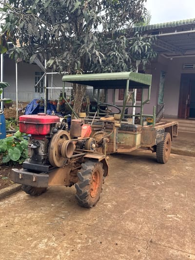 An aged, custom-built two-wheel drive agricultural vehicle, commonly referred to as a power tiller tractor with an integrated flatbed trailer, is parked on a dirt-stained concrete or packed earth surface in Ko H'Neh, Vietnam. The vehicle's front section features a prominent red, single-cylinder diesel engine, an exposed belt-driven transmission system, and two large, deeply-treaded agricultural tires caked with mud. A black front bumper is visible. The central operating area includes a steering wheel and is covered by a utilitarian green metal-framed canopy. The rear consists of a low-sided flatbed trailer with smaller, smoother tires. The entire vehicle exhibits heavy wear, including rust, grime, and mud accumulation, indicative of extensive use. Small, dark, spherical debris, consistent with coffee beans, are scattered across the ground.
In the background, to the left of the vehicle, a multi-story white building with a covered porch and railings is partially visible, obscured by large-leafed green plants and a crumpled blue tarp covering indeterminate objects. Centered behind the vehicle, a large tree with dense, dark green foliage stands. To the right, a single-story house with a light brown/pinkish facade, an open double doorway, and a metal-framed roofing structure is present. Several potted plants hang near the right-side house. The sky is overcast. No individuals are present.