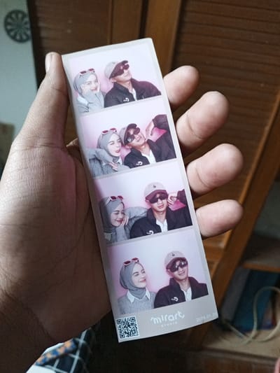 A left hand, displaying visible fingerprints and moderate skin tone, holds a vertical photo strip. The strip features four identical square frames, each depicting a female on the left and a male on the right against a solid light pink background. The female wears a light gray hijab, a white collared shirt, and a gray knitted sweater, with red-framed cat-eye sunglasses pushed onto her hijab. The male wears a beige baseball cap, dark round sunglasses, a white shirt, and a dark jacket.

In the first frame, the female smiles slightly while the male looks upward to the right with a faint smile. The second frame shows the female playfully poking out her tongue with her left arm resting on the male's right shoulder, who makes a similar playful expression. The third frame depicts the female smiling with her head resting on the male's right shoulder, who looks forward. In the fourth frame, both individuals smile while looking towards the left side of the frame.

The bottom section of the photo strip includes a QR code on the left, the "m!rart STUDIO" logo in the center, and the date "2025.01.20" on the right. The background of the primary image features vertical wooden paneling on the right and bottom, and a faint, spoked circular object on a lighter wall to the upper left. The background environment does not contain identifiable features that specify a location within the city of Al Haouafate, Morocco.