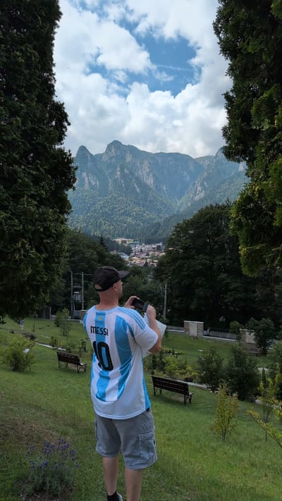 A man wearing a white and light blue striped jersey with "MESSI 10" on the back stands in a grassy area, holding a phone in his hands as if taking a picture. He is facing away from the camera and towards a mountainous landscape. In the foreground, there is a small lavender plant. In the background, there are trees, benches, and a town nestled at the base of the mountains. The sky is cloudy and blue. The overall scene suggests a tourist enjoying the scenery in Bușteni, Romania.