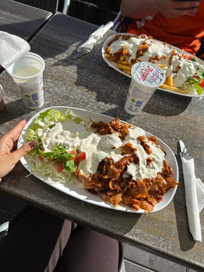 The image shows two plates of Döner meat, topped with a white sauce, served on a bed of shredded lettuce and tomatoes, along with a side of french fries on one of the plates. Two containers of Ayran are present. One plate is being held by a hand with polished nails. The setting appears to be an outdoor table. Given the presence of Ayran and Döner, the location could be a restaurant in Bruchköbel, Germany.