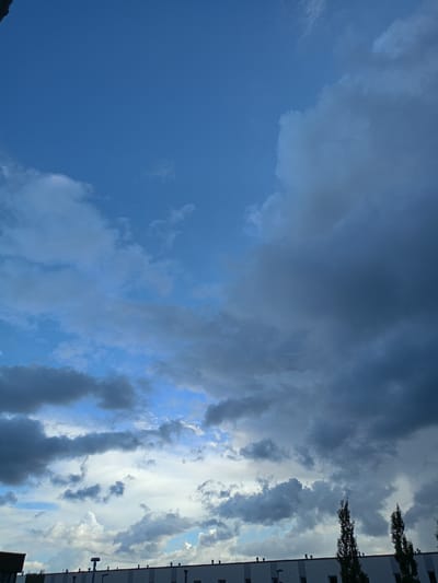 An exterior, low-angle shot captures a cloudy sky above buildings and trees in Peetri alevik, Estonia. The sky is a mix of deep blue and dark gray clouds, with some patches of bright white clouds. A long, white building with a dark roof is visible across the bottom of the frame, with a line of trees in front of it. The overall impression is of a stormy day.