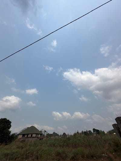 The image displays an outdoor scene in Agbogugu, Nigeria, under a partly cloudy sky. The upper two-thirds of the frame are dominated by a pale blue sky with scattered white cumulus clouds of varying sizes; some darker grey clouds are present towards the upper right. A single dark, thin line, likely an overhead utility wire, traverses diagonally from the upper left corner towards the mid-right of the sky.
The lower third of the frame shows an overgrown field or vacant lot. The foreground consists of dry, tall, wild grass and weeds, predominantly in shades of light green and brown. In the midground, towards the left, a partially constructed or unfinished building with a dark green, multi-faceted pyramidal roof is visible. Its walls are exposed concrete or blockwork, and window openings are present. Further into the background, several other structures with various roof types, including some with darker, possibly corrugated metal or tiled roofs, are discernible amidst sparse trees and vegetation. On the far right edge of the frame, a concrete structure, possibly a pillar or incomplete wall section, with exposed rebar at its top, is present. No individuals, specific actions, or interactions are visible within the scene.