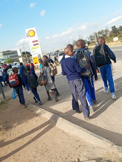 A group of approximately 10-15 young individuals, appearing to be students, are depicted walking on an unpaved dirt sidewalk adjacent to a paved multi-lane road in Ildamat ward, Kenya. Most individuals are dressed in dark blue uniforms, consisting of long-sleeved tops and trousers; one individual wears a plaid dress. Multiple backpacks of varying colors (red, black, grey, beige) are carried on their backs, and one person holds a large, clear plastic water bottle.

In the mid-ground on the left, a prominent Shell gas station pylon sign stands, displaying the red and yellow Shell logo at its apex, digital fuel prices (e.g., 195.99, 189.99), and text including "FuelSave," "Helix," and "Way In" with an arrow. The sky is clear blue with scattered white clouds. Long shadows are cast on the dirt sidewalk from the individuals, indicating a low sun angle. The environment includes parked vehicles along the road, low-rise buildings in the left background, and sparse vegetation in the distant background.