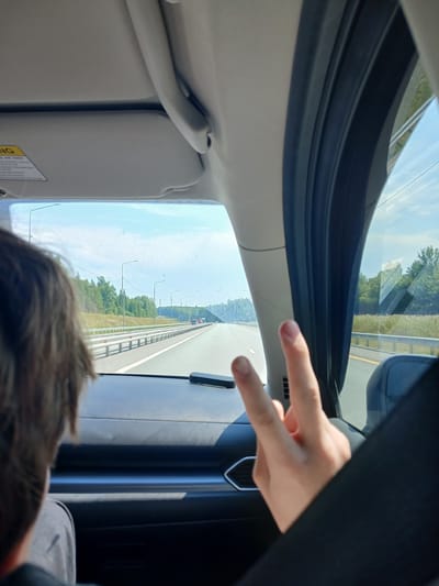 The image is taken from inside a vehicle, looking forward through the windshield. The foreground shows the back of a person's head and shoulder on the left, and a hand making a peace sign on the right. The vehicle appears to be on a highway with multiple lanes. The landscape outside consists of trees, fields, and a blue sky with some clouds. Streetlights are visible along the side of the road. There are other vehicles in the distance. The dashboard and sun visor of the car are also visible. The scene is set on a sunny day. The provided hint about Novye Koshelei, Russia, suggests this highway location is in or near that area.