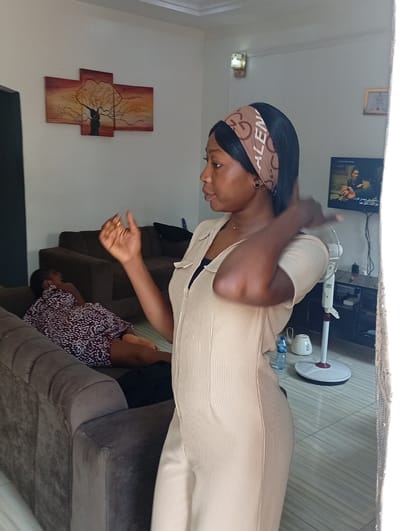 The image depicts a woman standing indoors, likely in a residential setting in Basawa, Nigeria. She is wearing a light brown jumpsuit and a headband with the word "Valentino" printed on it. Her right arm is raised. In the background, a child is lying on a brown sofa. Behind them, another brown sofa is visible. On the wall, there is an artwork depicting a tree. A television is also visible, displaying an individual on screen. A white electric fan, a water bottle, and an electric kettle are situated near the television stand. The room has light-colored walls and tiled floors. A curtain is partially visible on the right edge of the image.