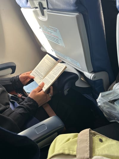 A person is seated on an airplane, reading a book. The book appears to be religious text. The person is wearing a black long-sleeved garment with lace cuffs and a silver ring on their right hand. A smartphone is placed on top of the book. The environment consists of airplane seats, with a tray table visible in the background. The lighting suggests it is daytime.