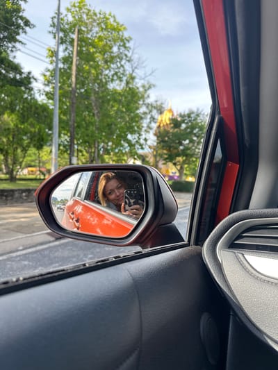 In Ao Luek, Thailand, a person is captured in a rearview mirror selfie from inside a vehicle. The person is smiling and holding a smartphone to take the photo. The mirror reflects the interior of the orange-colored car and the person's face. Outside the car, trees and a golden-roofed structure, possibly a temple, are visible. The scene suggests a travel or leisure context.