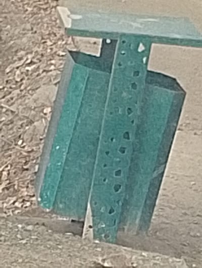The scene depicts an outdoor environment in the city of Qanlikul, Uzbekistan, featuring a teal-colored metal structural component. This component consists of a horizontal top section, presenting as a flat, broad surface. Directly below it, a vertical support column made of perforated teal metal is visible, exhibiting multiple small, distinct square and circular holes arranged in a vertical pattern. To the left of this perforated column, a solid, flat vertical teal metal panel forms an additional part of the structure. The base of the metal structure rests on an uneven ground surface composed of light-colored earth, possibly sand or packed dirt, with scattered small organic debris or gravel. No individuals, animals, or active events are present in the frame. The illumination suggests natural daylight.
