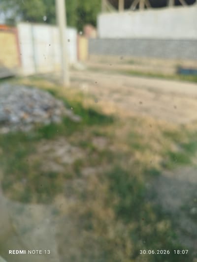 This is a slightly blurry image taken outdoors, likely in a rural or semi-rural setting in Асан-Чек, Kyrgyzstan. The foreground is dominated by dried and green grass, with some scattered stones or debris on the left. In the background, there are structures that appear to be under construction, with wooden beams visible against a pale wall. A white post stands vertically near the center of the image. The overall scene suggests a sunny day, as indicated by the lighting.

Visible text overlays in the bottom left corner read "REDMI NOTE 13", and in the bottom right corner, "30.06.2026 18:07". These indicate the device used to take the photo and the date and time it was captured. The blurriness may be due to the camera lens or the movement of the photographer. No people are visible in the image.