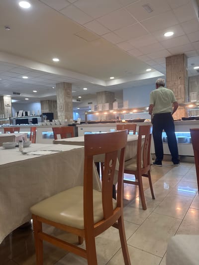The image shows a man, appearing to be a tourist, in a buffet-style restaurant. He is standing in front of a food serving station, looking at the food options. The restaurant is furnished with tables and chairs, and the lighting suggests it is daytime. The floor is tiled. The man is wearing a light green short-sleeved shirt and dark pants. The overall scene suggests a typical dining experience in a restaurant. Fuengirola, Spain is a popular tourist destination known for its beaches and restaurants, and this scene is consistent with what one might find in such a location.