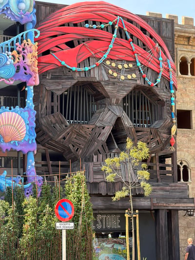 The image shows the exterior of a building designed to resemble a giant pirate skull in Santa Ponça, Spain. The skull structure is made of weathered wooden planks and features large openings for eyes, covered by vertical bars. A red, cloth-like material wraps around the top of the skull, resembling a turban, adorned with blue and gold beaded necklaces and chains with gold coin-like discs. To the left of the skull is a brightly colored, abstract sculpture in shades of blue, purple, and pink, with seashell and coral-like elements. In front of the building, there is a small, leafy tree and dense green bushes. A "no stopping" traffic sign with the text "EXCEPTO BUS REGULAR" (Except Regular Bus) is visible in the foreground. A sign with the word "STORE" in illuminated lettering is mounted below the skull. In the background, part of an adjacent building with arched windows and stone facade is visible. A man is standing on the right side of the frame, facing away from the camera. The sky is overcast.