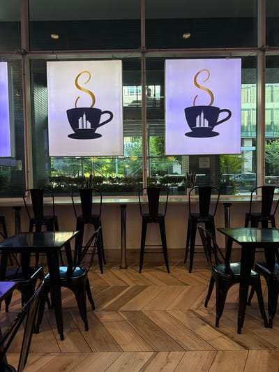 The image depicts the interior of a cafe or restaurant, possibly in Weehawken, United States of America. The scene is dominated by a series of tables and chairs. The chairs are modern, black metal stools, arranged around metal-topped tables. The flooring is a distinctive herringbone pattern made of light-colored wood planks.
Two large, illuminated signs are prominently displayed. Each sign features a stylized graphic of a coffee cup silhouette with a golden, swirling steam trail. Within the cup's silhouette, a city skyline is depicted with several rectangular buildings. The signs are mounted on a wall, partially obscured by the reflection of outdoor greenery and buildings in the large windows behind them.
Through the windows, an urban or suburban setting is visible, with trees and buildings suggesting an outdoor environment. The lighting within the establishment appears to be a mix of artificial light from the signs and natural light filtering through the windows. The overall atmosphere seems calm and unoccupied, with no people visible in the immediate seating area. The time of day is indeterminate, but the artificial lighting suggests it could be evening or a dimly lit interior. A small, round object, possibly a coin, is visible on a surface to the left of the upper sign.