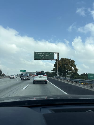 The image captures a view from a vehicle on a highway in Long Beach, California. A large green overhead sign indicates the Cherry Ave South/Signal Hill exit is 1 mile ahead. Other vehicles, including a white SUV, are visible in the adjacent lanes. The sky is partly cloudy. The road appears to be a multi-lane highway with a concrete barrier on the right side. Vegetation, including trees, can be seen beyond the barrier. The image is taken from within a vehicle, as evidenced by the car frame visible on the right side.