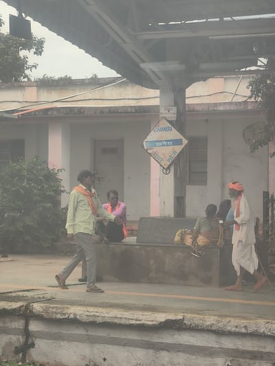 The image depicts a platform at the Chhanera railway station in India. Several individuals are present on the platform. In the foreground, a man wearing a light-colored, patterned shirt, grey trousers, and an orange scarf walks towards the left. To his right, a man in a pink shirt and an orange scarf sits on a raised platform. Further to the right, an elderly man in a white outfit and an orange turban is serving a drink from a small bowl to a woman seated on the platform, who is wearing a yellow and floral patterned sari. Another woman is seated behind her. A diamond-shaped station sign indicating "CHANNERA" and "छनेरा" along with "WCR" (West Central Railway) is visible on a pole behind the seated individuals. The platform has a textured yellow line along its edge. The background shows a building with windows and a door, possibly a station building, with overhead structures and overhead wires. Trees and foliage are visible on the left side of the image. The overall scene suggests a typical day at an Indian railway station with people waiting or transiting.