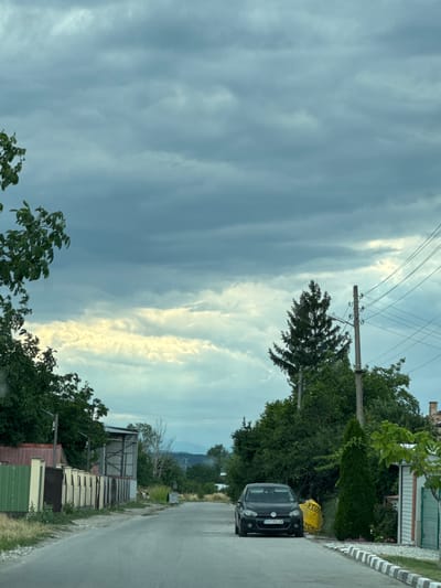 This is a vertical, outdoor photograph taken from a slightly elevated viewpoint, possibly from inside a vehicle. The scene depicts a rural road in Gelemenovo, Bulgaria, with buildings and lush greenery lining one side. The sky is a dominant feature, filled with dramatic, overcast clouds with streaks of bright sunlight breaking through in the distance, suggesting either the beginning or end of a storm, or perhaps a sunrise/sunset.
On the right side of the road, a dark-colored Volkswagen hatchback is parked. The car has a license plate visible, reading "PA 3956 KM". Next to the car, a bright yellow bag, possibly containing construction material, is visible with partial text that includes "YTONG" and "TONG". Further down the road, several simple, utilitarian structures with corrugated metal roofs are present. One structure appears to be an open-sided shed with metal shelving units inside. A power line pole with multiple wires stretches across the frame, adding to the rural atmosphere.
The vegetation is dense, with mature trees and bushes forming a green backdrop to the scene. In the far distance, hills or mountains are faintly visible under the cloudy sky. The overall mood is somewhat somber due to the overcast sky, but the patches of sunlight add a touch of hope or dramatic flair. The time of day is difficult to pinpoint precisely due to the cloud cover, but the quality of light suggests it might be late afternoon or early evening.