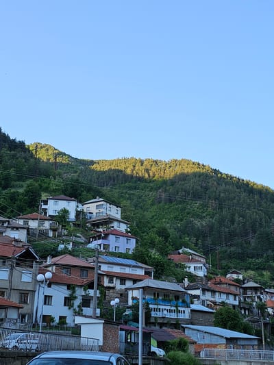 The image shows a residential area in Erma Reka, Bulgaria, built on a steep hillside. The houses have a variety of architectural styles, with red-tiled roofs being common. Trees and other vegetation surround the houses. Overhead power lines run through the area. The left side of the frame shows a clear, blue sky. The hillside extends from the center to the left, bathed in sunlight on the crest. A car is partially visible on the right.