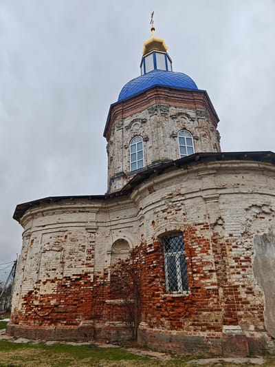 The image shows a low-angle shot of the Church of the Intercession of the Holy Virgin in the посёлок совхоза "Буденновец", Russia, under an overcast sky. The church is made of red brick covered with white plaster that is peeling in many places, revealing the brick underneath. The cylindrical part of the church has one visible barred window. Above this, the structure narrows and has two arched windows. On top of that is a blue dome, with a white and gold spire capped with a cross. The church appears to be in a state of disrepair. Ground with short grass visible at the bottom of the image. Bare branches from a small plant are at the base of the church. Power lines are visible on the left.