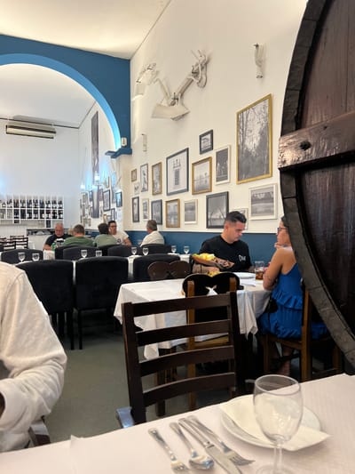 The image depicts the interior of a restaurant in Coruche, Portugal. The scene is a well-lit dining area with white walls and a distinct blue trim outlining an archway and a lower wall section. A collection of framed monochrome photographs of various sizes adorns the main wall, complemented by abstract white sculptural elements mounted higher up.
Multiple dining tables are present, each covered with a white tablecloth. Chairs are a mix of dark upholstered models and dark wooden styles. In the mid-ground, a man with short dark hair, wearing a black t-shirt with a white logo, is seated at a table opposite a woman with dark hair, wearing a blue tiered dress. A basket containing food, along with drinks in glasses, rests on their table. They appear to be engaged in conversation. Partially obscuring the right side of the frame is a large, dark-stained wooden barrel.
Further into the background, several other patrons, predominantly men, are seated at tables, some appearing to be conversing or dining. One man wears a green shirt, another a white shirt. On the far left in the background, a white shelving unit is stocked with numerous wine bottles. An air conditioning unit is mounted high on the wall near the archway. In the immediate foreground on the left, a portion of a person wearing a white long-sleeved shirt is visible. A table setting in the foreground displays silverware (fork, spoon, knife), a white plate, a white napkin, and an empty stemmed glass.