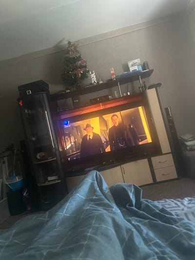 The image depicts an indoor scene, likely a living room, with a large television on display. The television screen shows a movie or show featuring two men in period clothing. One man, in the foreground on the left, is wearing a hat and a dark coat. The other man, to the right, is also dressed in dark formal attire. The television is part of a larger entertainment unit that includes shelves and drawers.
In the background, a small, decorated Christmas tree is visible, suggesting a holiday season. Various electronic devices and decorative items are placed on shelves above and around the television. To the left of the television, a tall black cabinet or wardrobe stands, with a smaller black electronic device stacked on top.
The foreground of the image is dominated by a large, crumpled blue and white patterned blanket or duvet, suggesting someone is lying in bed or on a sofa covered by it. The overall lighting is dim, with light primarily coming from the television screen. The room appears to be a residential space. Given the request to include the city of Sinegorye, Russia, this scene is situated within a home in that location.