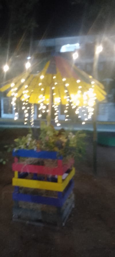 A blurry outdoor shot captures a colorful wooden planter box, topped with greenery. The box is stacked with layers painted in blue, red, and yellow. Above the planter, a yellow awning, strung with fairy lights, creates a soft glow. The image is taken at night, with dark surroundings and blurred background elements.