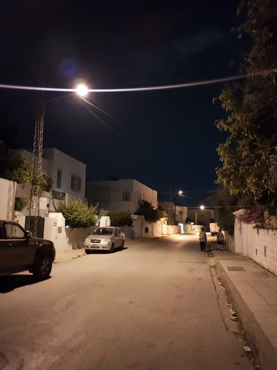 This nighttime image depicts a street scene in the city of Ariana, Tunisia. The sky is dark blue, with a bright street light illuminating a portion of the road and casting long shadows. A silver sedan is parked on the right side of the street, with its headlights off. A darker-colored SUV is parked on the left side of the street, with its headlights on. Two individuals are walking away from the camera in the distance, down the center of the street. White buildings with windows line both sides of the street, and lush green trees and purple flowers are visible on the right side. The street is paved, and a concrete sidewalk runs along the right side, with a drainage ditch alongside it. The overall impression is one of a quiet, residential street at night.
