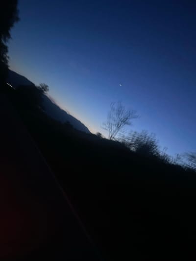 The image depicts a twilight scene with a deep blue sky and a sliver of a crescent moon visible. In the foreground, a dark silhouette of trees and a hillside dominates the lower portion of the frame. A faint, blurry light source, possibly from a distant vehicle or building, is visible on the left side of the image, near the base of the hillside. The overall impression is that of a natural landscape at dusk or dawn, with a hint of human presence. The image appears to be taken from a moving vehicle, given the slight blur and tilt. There are no people or identifiable structures visible in detail, only the broad strokes of a natural environment. Without further context or specific markers within the image, pinpointing an exact location within the city of Hvostiane, Bulgaria, is not possible. The scene is generic enough to be found in many rural or semi-rural areas.