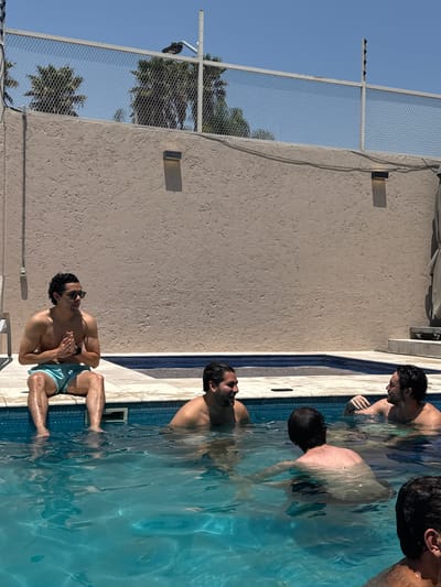 The image shows a group of men in a swimming pool. One man is sitting on the edge of the pool with his feet in the water, wearing sunglasses and swim trunks. The other men are in the pool, some partially submerged. The pool is bordered by a light-colored wall with two visible rectangular fixtures and a wire fence above it. Palm trees are visible behind the fence, and the sky is clear and blue. Based on the prompt, this scene is located in Santa Cruz Tehuixpango, Mexico.