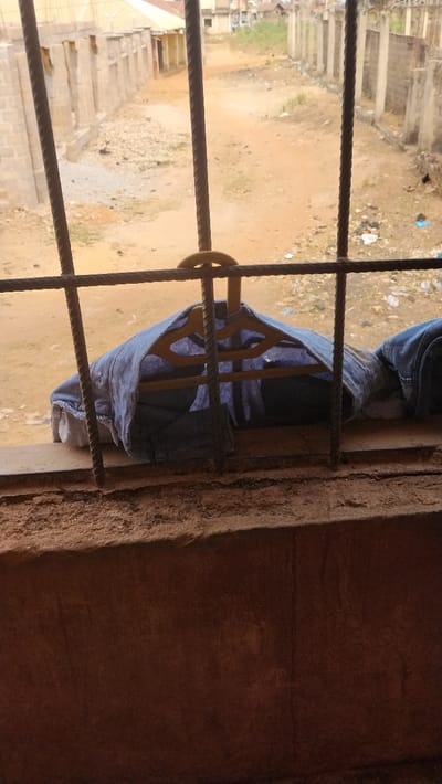 The scene depicts an exterior view through a window in Isieke, Nigeria. In the immediate foreground, a textured brown wall or window ledge is visible, covered in fine brown dust. Directly beyond this, a dark metal security grille, composed of vertical and horizontal rebar, obscures part of the view. A light blue denim jacket, draped over a yellow plastic clothes hanger, rests on the window ledge behind the grille, partially covering a white garment. Beyond the window structure, an unpaved, reddish-brown dirt path, uneven with scattered gravel and refuse, extends into the distance. Along the left and right sides of this path, multiple incomplete or dilapidated single-story concrete block structures are present. Further in the background, additional buildings with visible roofs are discernible under an overcast sky. No individuals are present in the frame.