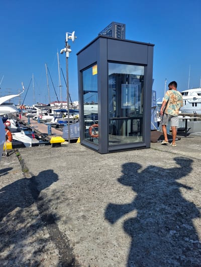 A man wearing a floral print shirt and grey shorts stands on a concrete pier, casting a large shadow. He is looking down at something beyond the frame. Beside him is a modern, grey kiosk with large glass windows, reflecting the surrounding environment. The kiosk has a metal grille structure on its left side.
The scene is a marina, with numerous sailboats docked in the background under a clear, bright blue sky, suggesting a sunny day. The water appears calm. Visible on the boats are life preservers. One of the boats has a Russian flag.
On the pier, near the kiosk, a yellow float is present. The concrete surface shows some wear and tear, with patches of darker discoloration. The shadows are sharp, indicating it is likely midday. The overall atmosphere appears relaxed and leisurely, characteristic of a marina on a pleasant day.
There is some visible text on the kiosk's glass, though it is mostly obscured or reflected. Partially visible text reads "D MAR" and "SOC". The context of Guzeripl', Russia, points to a marina in that region.