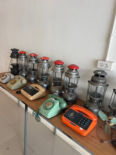 This media file showcases a collection of vintage items, primarily old rotary dial telephones and kerosene lanterns, arranged on a wooden surface against a textured white wall. Several kerosene lanterns, mostly made of metal with glass enclosures and some featuring red caps, are stacked vertically. Next to them, a row of telephones in various colors, including orange, light green, beige, and gray, are displayed. The orange telephone is a push-button model, while the others are rotary dial. A vintage electric fan with green blades is visible at the top of the image. A white electrical conduit and a power outlet are also present on the wall. The overall setting appears to be a storage area or a display of antique items. No people or specific activities are depicted. The lighting suggests it is daytime. The image has a slightly tilted perspective. Visible text includes price tags on some items, such as "560" and "131/152" on the orange phone, and "151/152" on one of the lanterns. The location context of Tambon Phraek Ha, Thailand, suggests a local setting for these items.